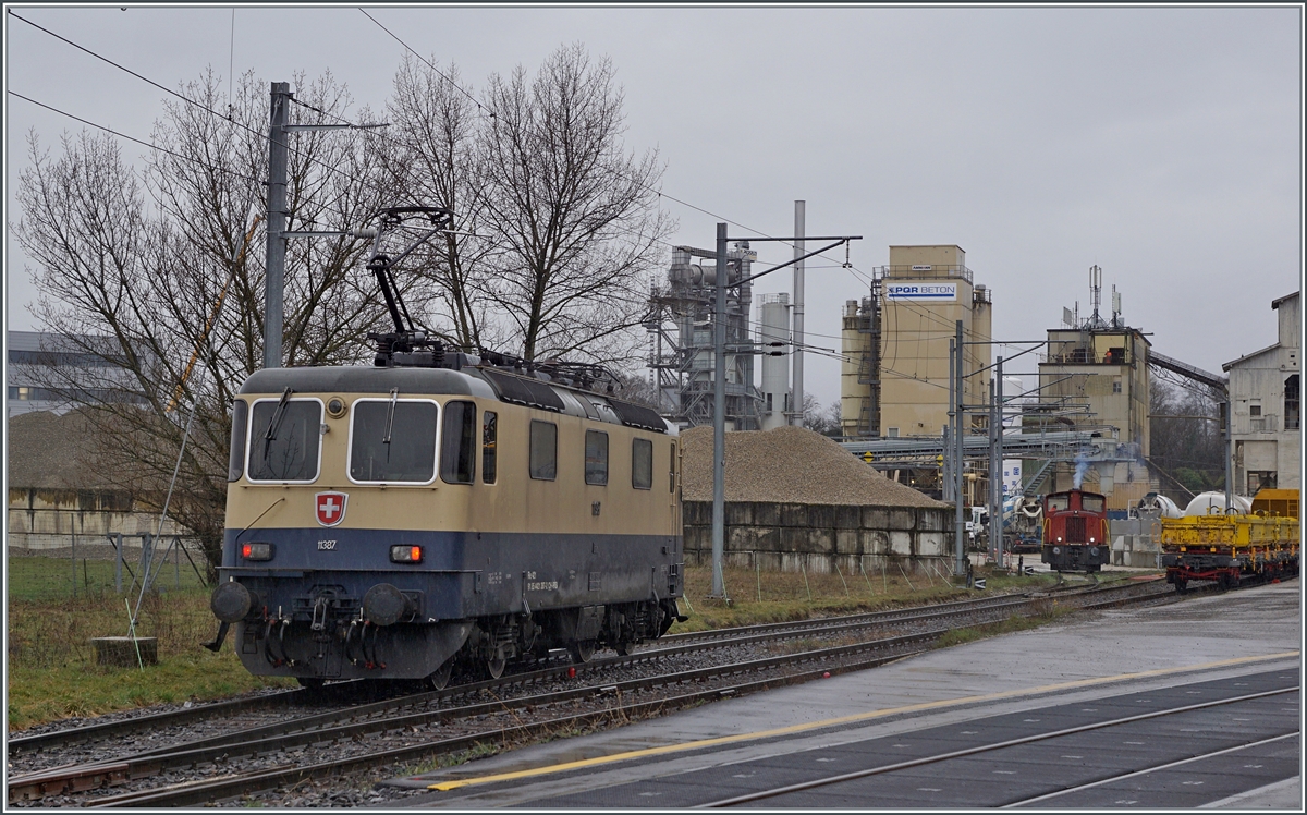 The IRSI/IGE  Rheingold  Re 4/4 II 11387 (Re 421 387-2), which came from Morges, leaves the route to Genève in Gland and travels over an industrial track towards the gravel works to pick up its train to Apples.
In the background you can see the BAM MBC Tm 232, clearing the way for the Re 4/4 II 11387.
The rented IRSI/IGE Re 4/4 II 11387 (Re 421 387-2) replaces the BAM MBC MBC Re 420 506, which is currently undergoing revision in Bellinzona. 

February 22, 2024