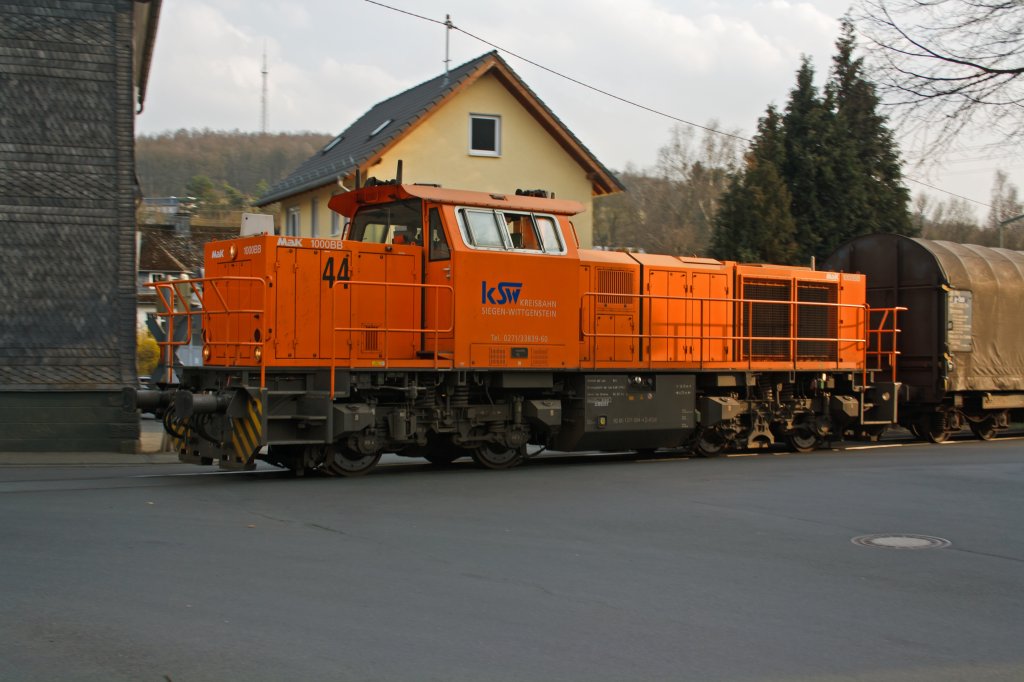 Locomotive 44 (MaK G 1000 BB) of the Kreisbahn Siegen-Wittgenstein (KSW) runs on KSW own rail, with a full goods train on 25/03/2011 at Neunkirchen over the railway crossing Wiesenstrae, towards  Salchendorf.