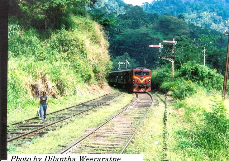 Unidentified class M5 locomotive is approaching Balana station bound to Colombo. The inner and outer semaphore home signals are visible in the picture. Note – village girl is using the rail track as a foot path due to lack of road ways.