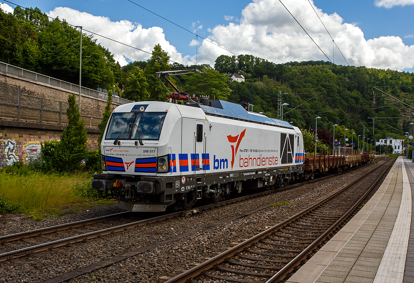El SIEMENS Vectron Dual Mode 248 077 (90 80 2248 077-0 D-NRAIL) de BM-Bahndienste GmbH (Karlsruhe) circula el 9 de junio de 2026 con un tren de plataformas (4 plataformas con bogies del tipo Rens), a través de la estación de Kirchen/Sieg hacia Siegen.

El SIEMENS Vectron DM fue fabricado en 2023 por Siemens Mobility GmbH en Múnich-Allach bajo el número de fábrica 23425 y entregado en enero de 2024 a BM-Bahndienste GmbH, que arrendó la locomotora a través de RRL - RIVE Rail Leasing Limited (Dublín), y la locomotora fue puesta en servicio por northrail GmbH.

La locomotora híbrida (Vectron Dual Mode) puede obtener su potencia de tracción de la catenaria aérea (2.400 kW de potencia en rueda) o generarla mediante un motor diésel MTU 16V 4000 R84 de 16 cilindros en V (2.400 kW de potencia del motor, 2.000 kW de potencia en rueda).